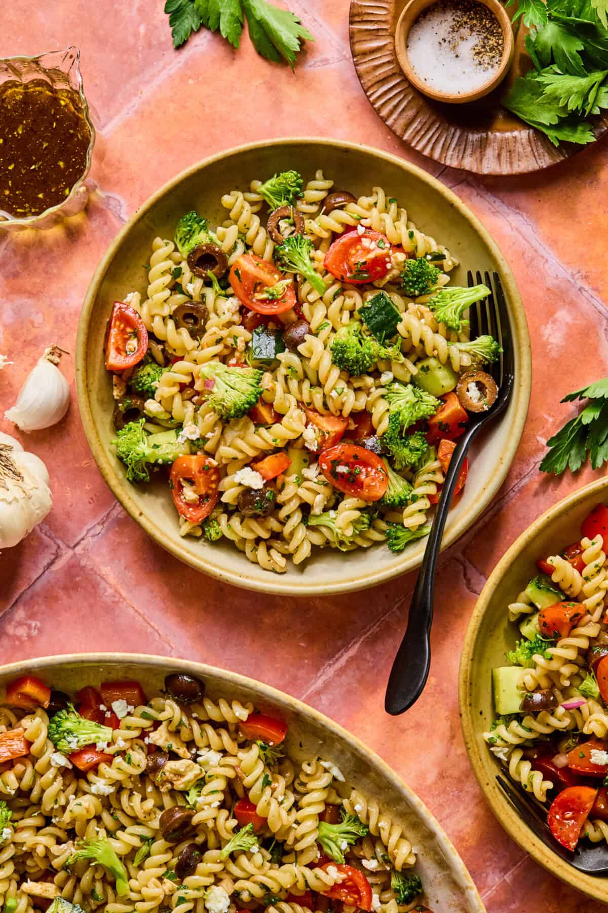 A plate of high protein pasta salad with cherry tomatoes, broccoli, olives, and herbs on a peach-colored surface, surrounded by extra plates of salad, fresh parsley, garlic, and a cup of dressing. A black fork rests on the main plate.