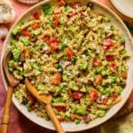 A large bowl of high protein pasta salad with rotini, cherry tomatoes, broccoli, red onion, olives, and herbs tossed in vinaigrette. Wooden serving utensils rest in the bowl, surrounded by plates and seasonings.