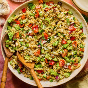 A large bowl of high protein pasta salad with rotini, cherry tomatoes, broccoli, red onion, olives, and herbs tossed in vinaigrette. Wooden serving utensils rest in the bowl, surrounded by plates and seasonings.