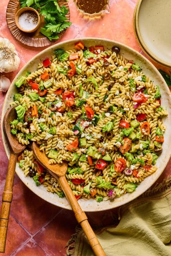A large bowl of high protein pasta salad with rotini, cherry tomatoes, broccoli, red onion, olives, and herbs tossed in vinaigrette. Wooden serving utensils rest in the bowl, surrounded by plates and seasonings.