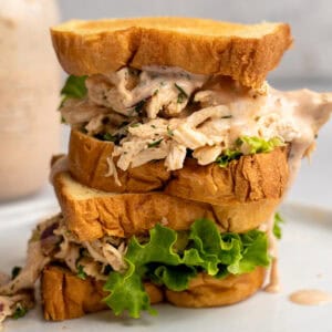 A tall sandwich filled with shredded Buffalo Ranch chicken salad, fresh green lettuce, and creamy dressing is stacked on a white plate. The sandwich is made with thick slices of toasted bread, and a jar of sauce is blurred in the background.