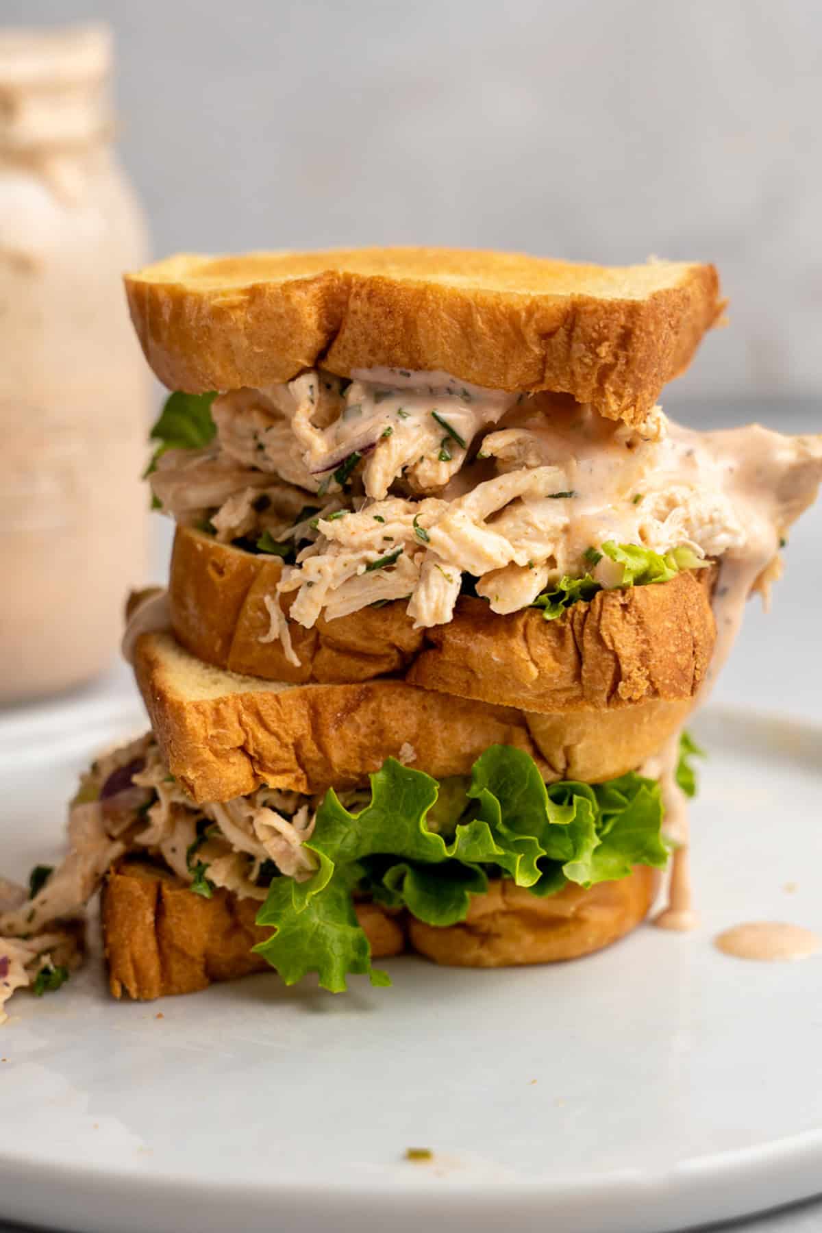 A tall sandwich filled with shredded Buffalo Ranch chicken salad, fresh green lettuce, and creamy dressing is stacked on a white plate. The sandwich is made with thick slices of toasted bread, and a jar of sauce is blurred in the background.