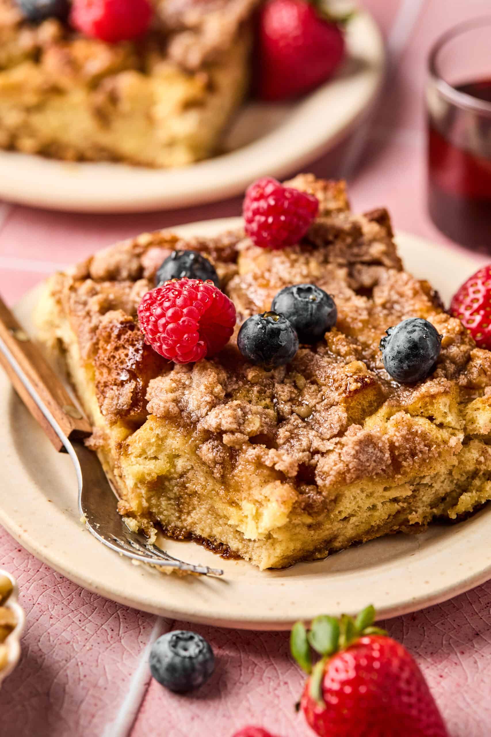 A close-up of a slice of crumbly coffee cake topped with fresh raspberries and blueberries, served on a beige plate, evokes the warmth of brioche French toast casserole. A fork rests beside the cake, with another serving and scattered berries visible in the background.