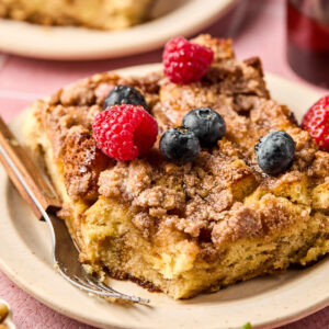 A close-up of a crumbly cake slice reminiscent of a Brioche French Toast Casserole, adorned with fresh raspberries and blueberries on a beige plate. The background features another slice, extra berries, and a glass of dark red liquid on the pink surface.