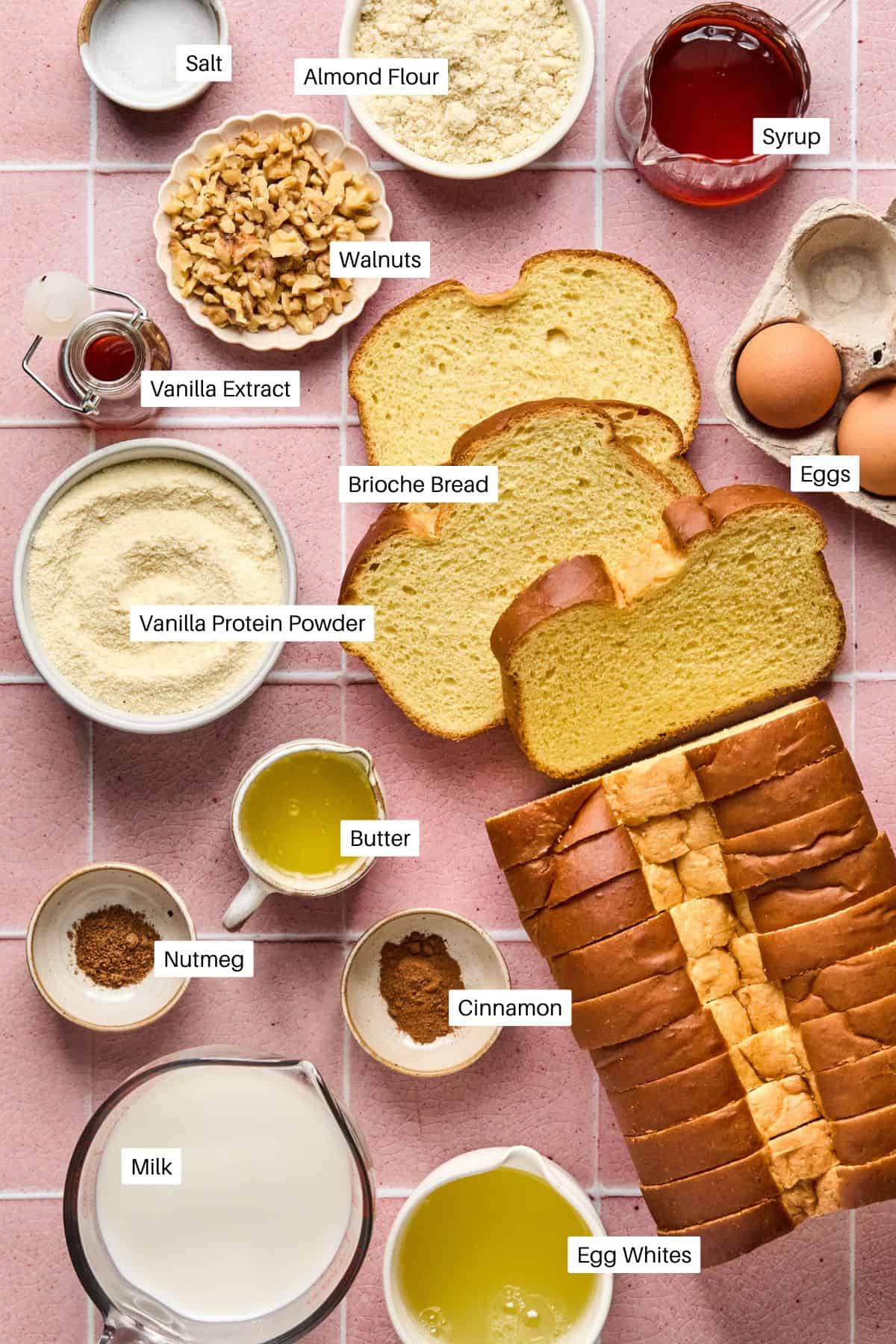 A top-down view of ingredients for a Brioche French Toast Casserole on a pink tiled surface. Includes salt, almond flour, walnuts, vanilla extract, syrup, eggs, vanilla protein powder, butter, nutmeg, cinnamon, milk, and egg whites. Perfect for a delightful breakfast twist!