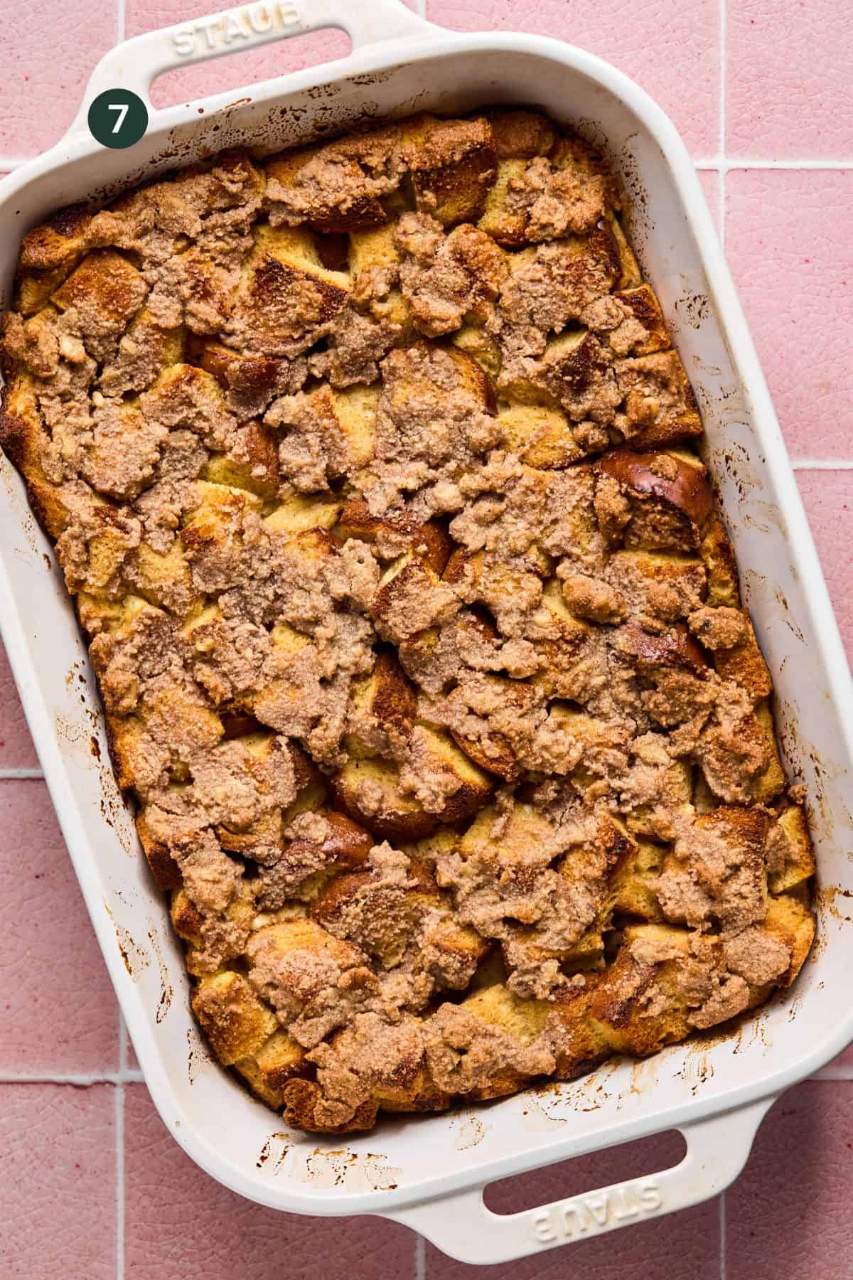 A baking dish filled with golden-brown Brioche French Toast Casserole, topped with a crumbly cinnamon streusel, rests on a pink tiled surface.