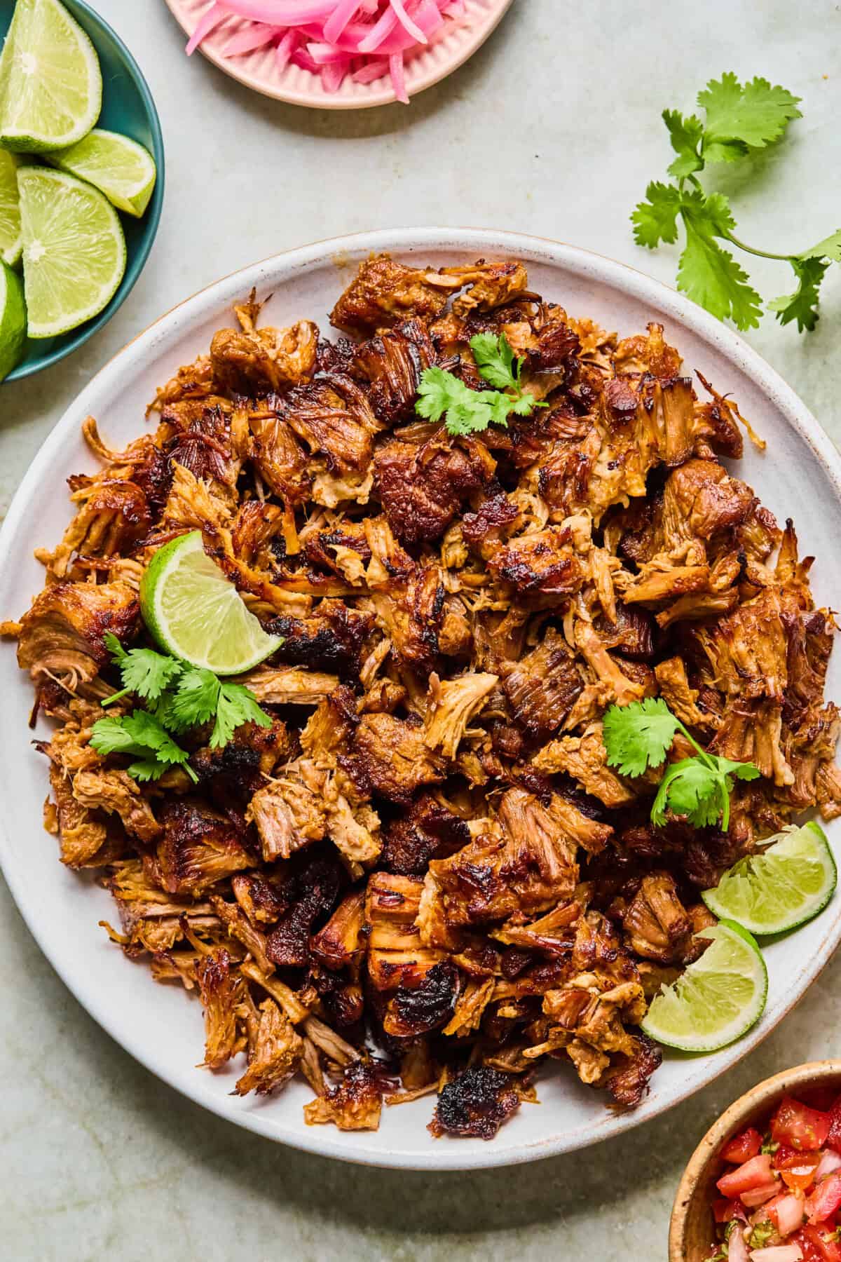 A plate of crispy, browned Instant Pot Carnitas topped with cilantro and lime wedges, surrounded by small bowls of lime slices, pickled onions, and fresh cilantro on a light surface.
