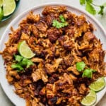 A plate of crispy, browned Instant Pot Carnitas topped with cilantro and lime wedges, surrounded by small bowls of lime slices, pickled onions, and fresh cilantro on a light surface.