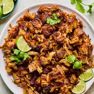 A plate of crispy, browned Instant Pot Carnitas topped with cilantro and lime wedges, surrounded by small bowls of lime slices, pickled onions, and fresh cilantro on a light surface.