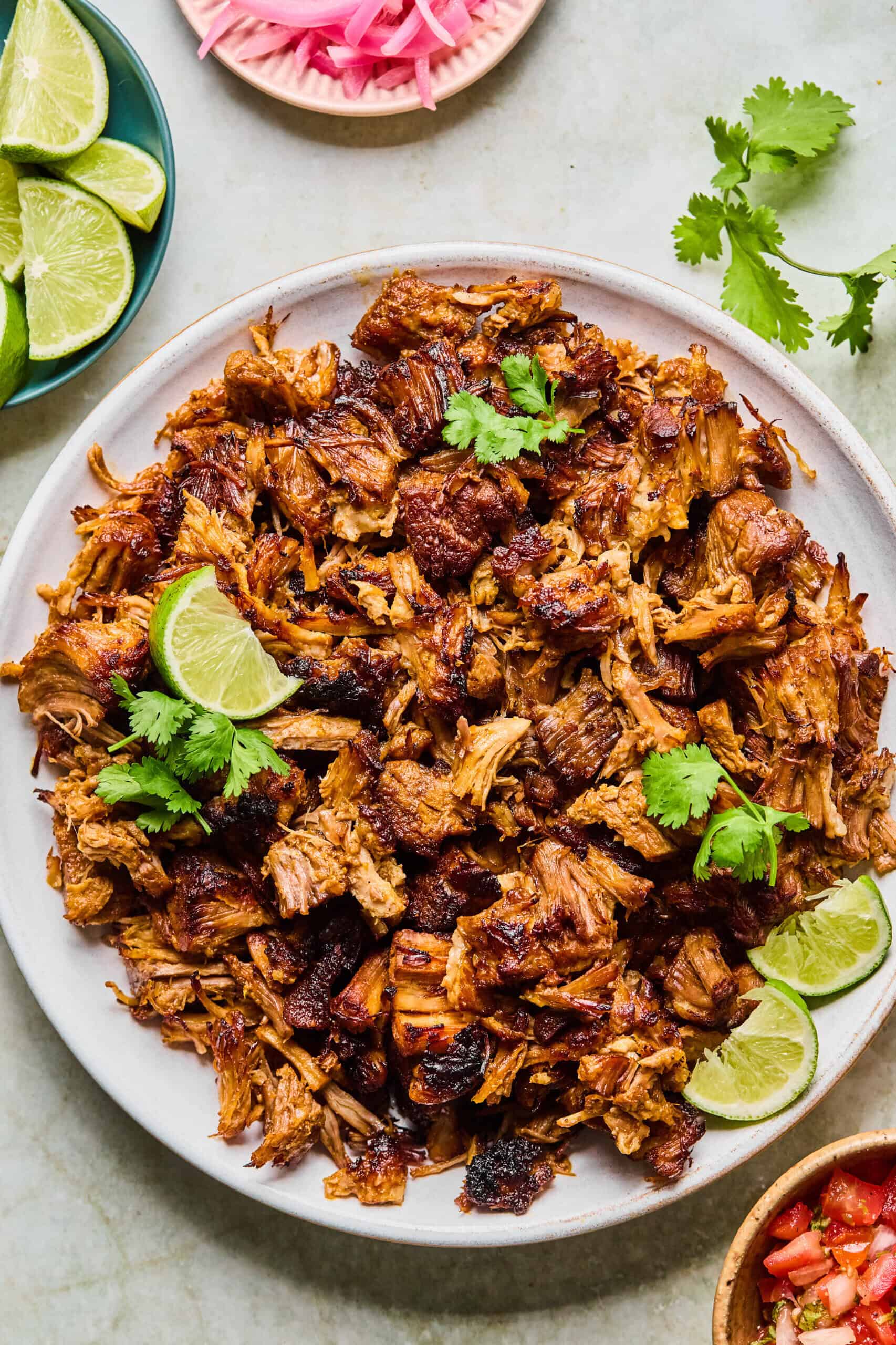 A plate of crispy, browned Instant Pot Carnitas topped with cilantro and lime wedges, surrounded by small bowls of lime slices, pickled onions, and fresh cilantro on a light surface.