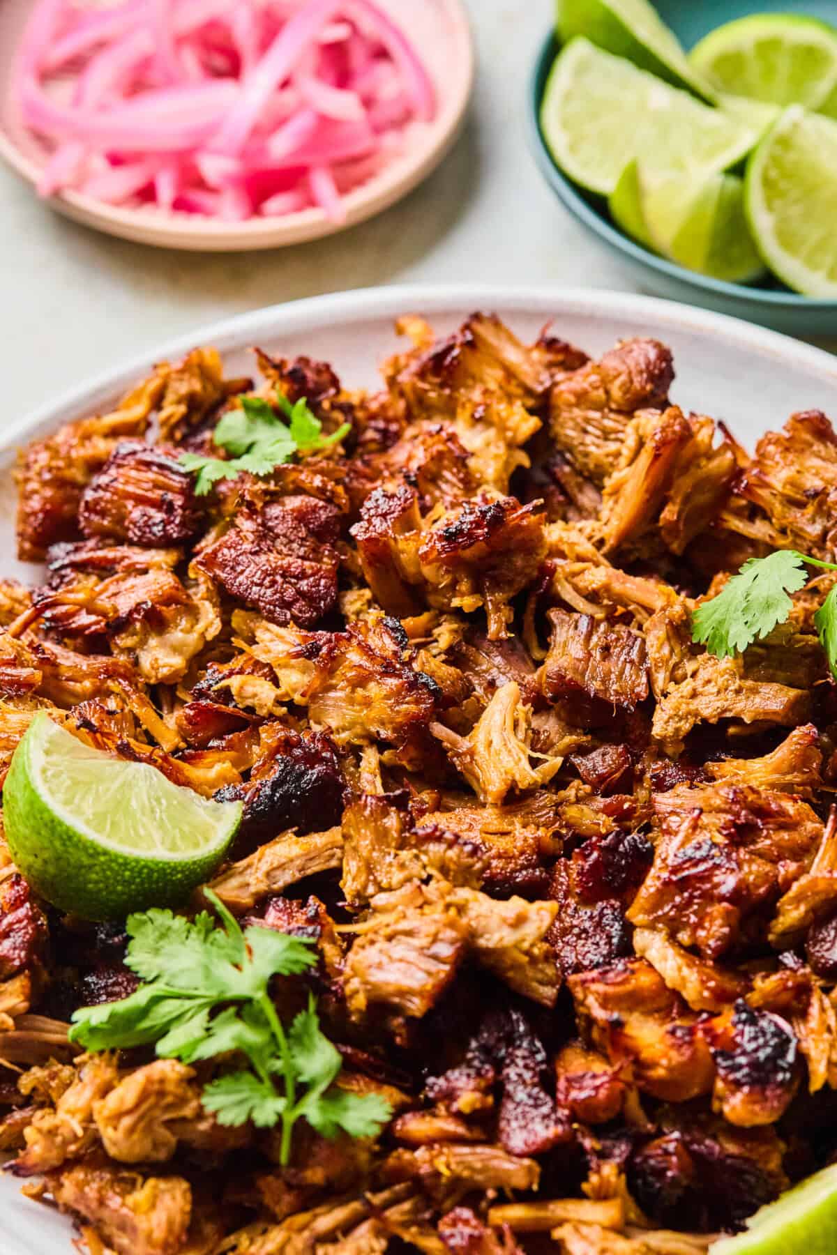 A plate of crispy, shredded Instant Pot Carnitas garnished with cilantro and lime wedges, with a bowl of lime slices and a dish of pickled red onions in the background.