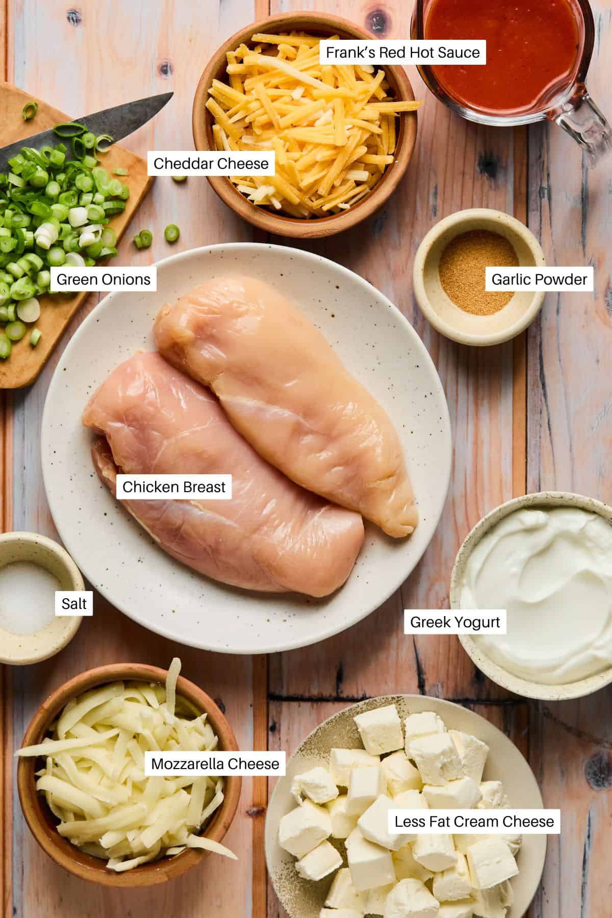 A flat lay of ingredients on a wooden surface hints at a delicious crock pot buffalo chicken dip, featuring raw chicken breasts, cheddar and mozzarella cheeses, green onions, Greek yogurt, less fat cream cheese, garlic powder, salt, and Franks Red Hot sauce.