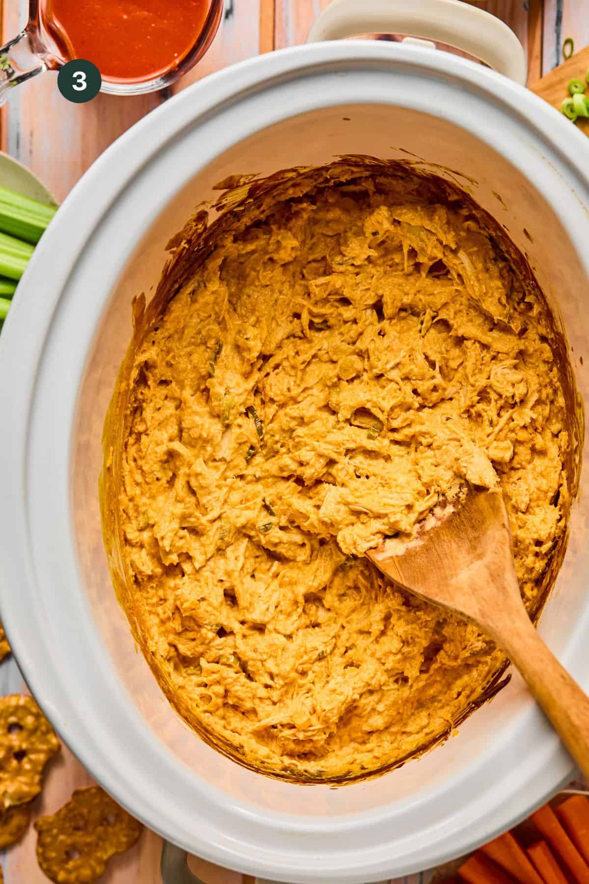 A ceramic crock pot brims with creamy buffalo chicken dip, stirred artfully with a wooden spoon. Nearby are crisp celery and carrot sticks, along with crunchy pretzels. In the background, a small bowl of hot sauce awaits for an extra kick.