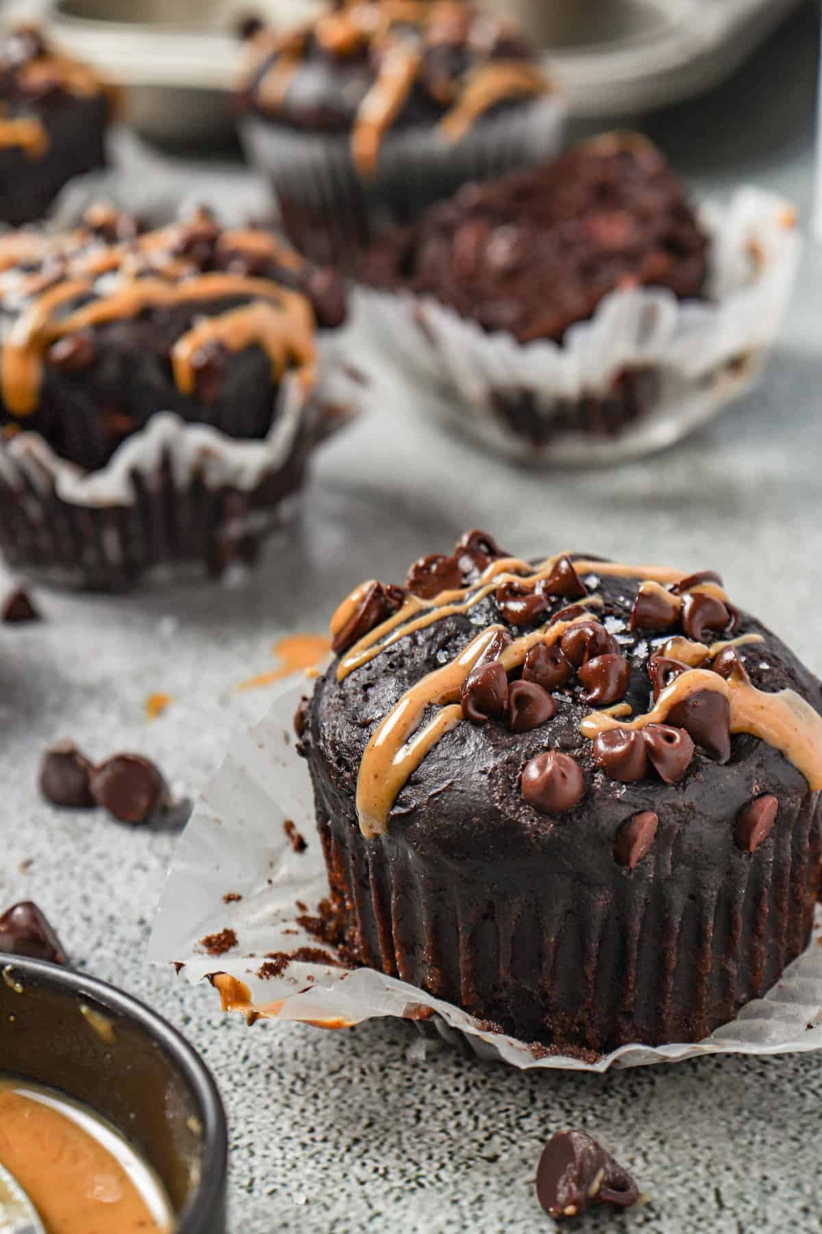Chocolate muffins with peanut butter drizzles on top with the muffin liner pulled down around the edges. 