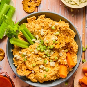 A delicious bowl of buffalo chickpea dip garnished with sliced green onions and celery sticks, surrounded by pretzel chips, carrot sticks, and blue cheese crumbs on a wooden table. A small jug of buffalo sauce is nearby—perfect for any crock pot buffalo chicken dip enthusiast seeking new flavors.