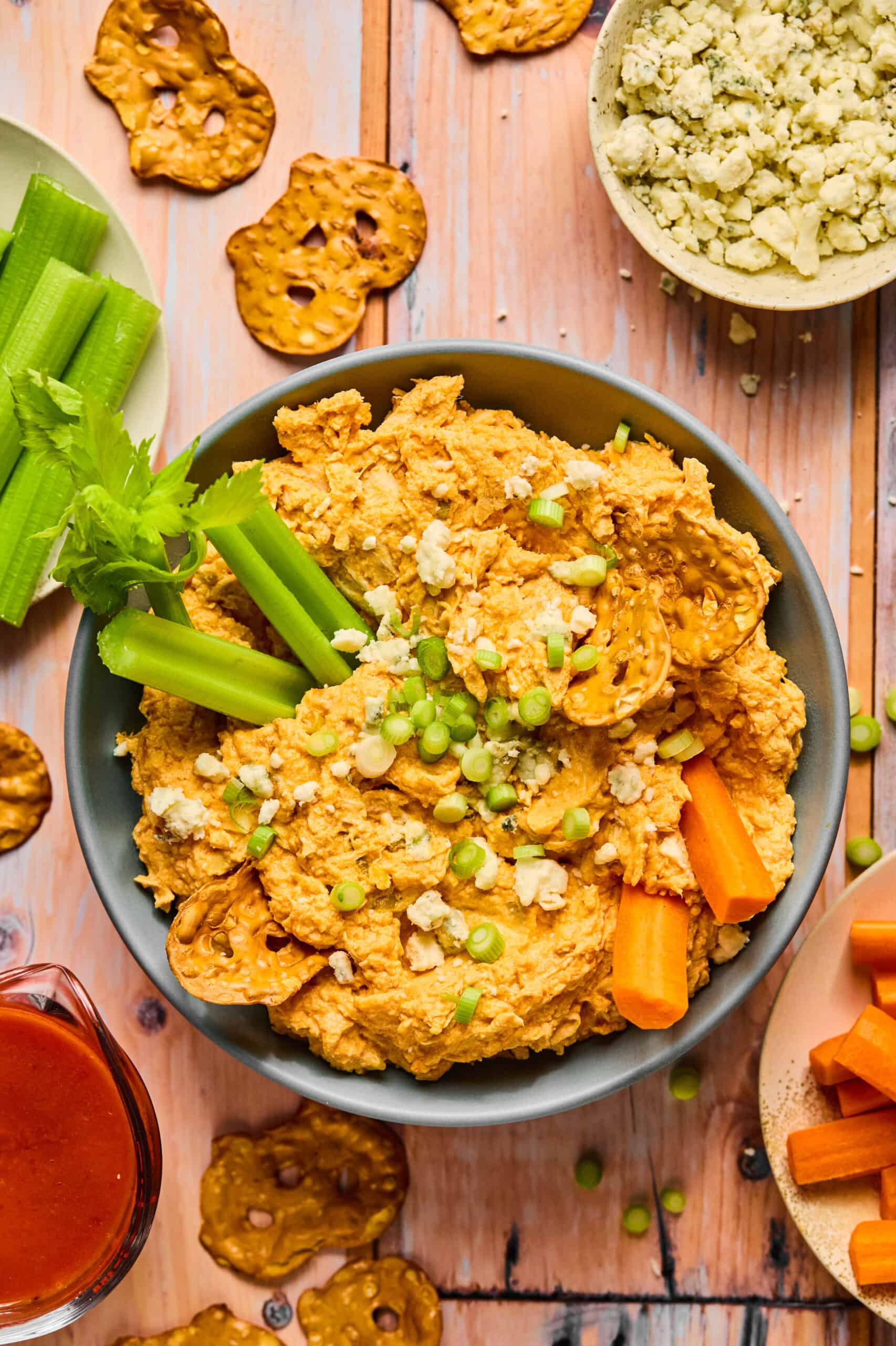 A delicious bowl of buffalo chickpea dip garnished with sliced green onions and celery sticks, surrounded by pretzel chips, carrot sticks, and blue cheese crumbs on a wooden table. A small jug of buffalo sauce is nearby—perfect for any crock pot buffalo chicken dip enthusiast seeking new flavors.