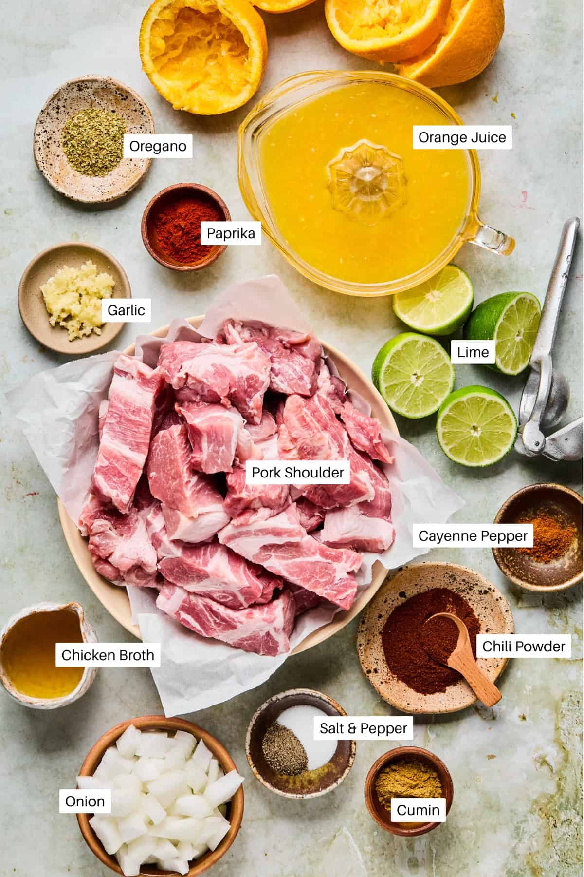 A flat lay of Instant Pot Carnitas ingredients, featuring raw pork shoulder, orange juice, lime, chicken broth, garlic, onion, and small bowls of oregano, paprika, cayenne pepper, chili powder, salt and pepper, and cumin.