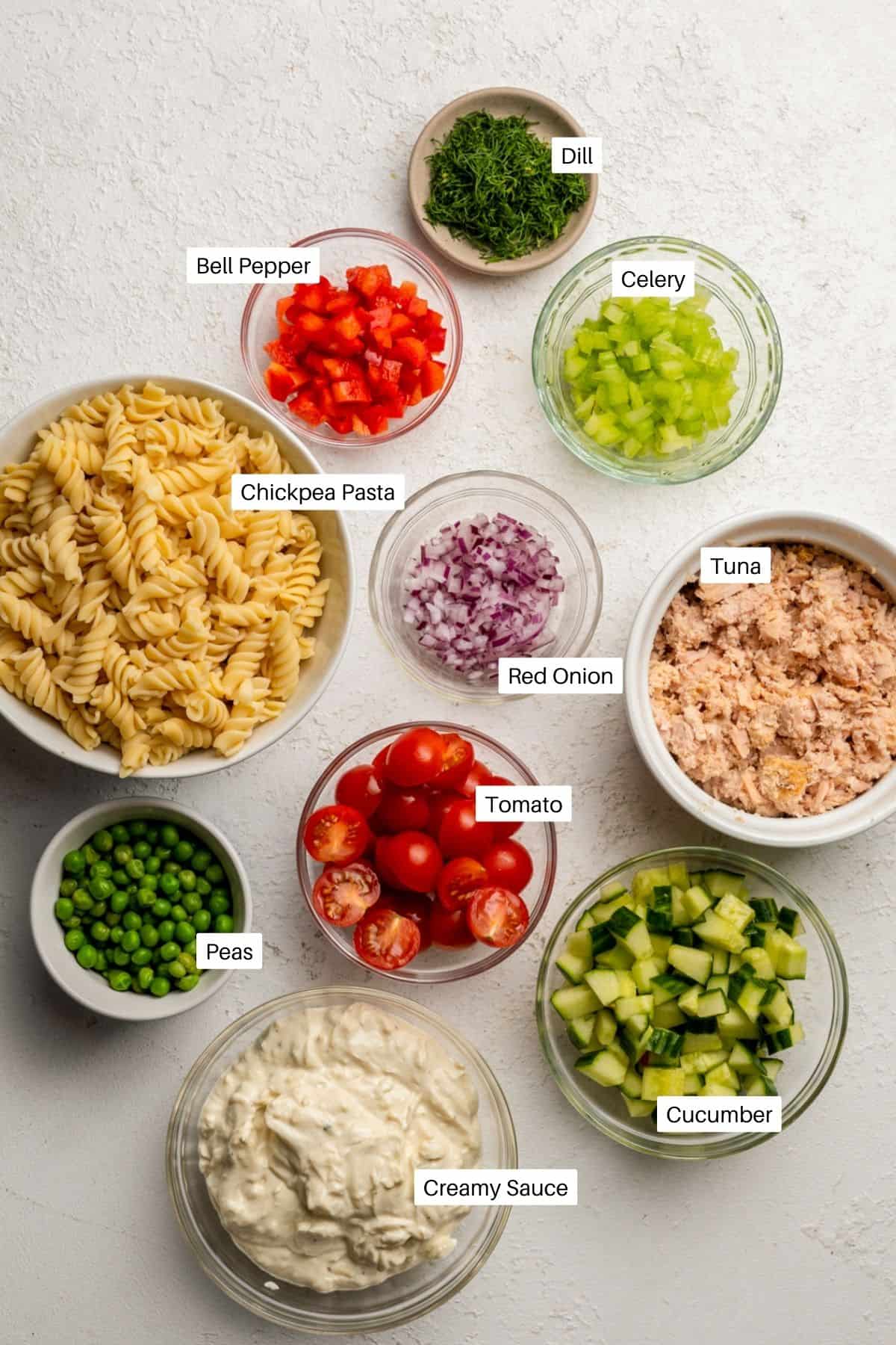 Dill, bell pepper, celery, chickpea pasta, red onion, canned tuna drained, tomtato, cucumber, peas and a creamy sauce for the salad laid out. 