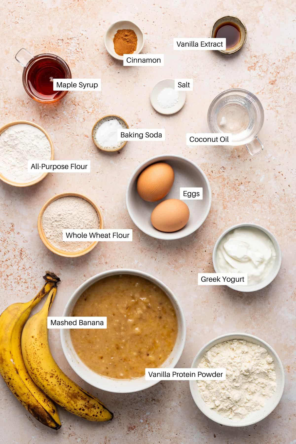 Various labeled ingredients for Protein Banana Bread are arranged on a textured surface, including bananas, mashed banana, vanilla protein powder, Greek yogurt, eggs, coconut oil, flour, maple syrup, and spices.