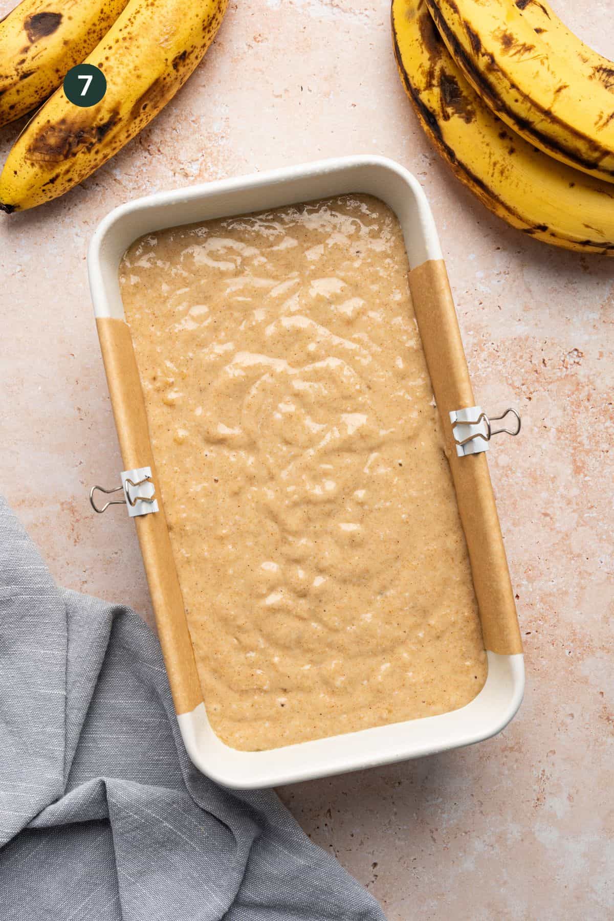 A loaf pan filled with protein banana bread batter, ready to be baked. The pan is lined with parchment paper and secured with metal clips. Ripe bananas are in the top corner, and a gray cloth is draped nearby on a textured surface.