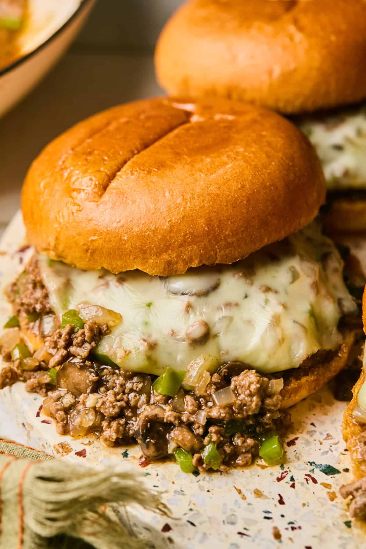 Close-up of a sandwich with a grilled beef patty topped with melted cheese, chopped green peppers, and onions, reminiscent of a Philly Cheesesteak, all served in a toasted bun. The sandwich rests on speckled paper, echoing the sloppy indulgence of Sloppy Joes, with another sandwich partially visible behind.