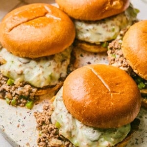 Four burgers with melted cheese and seasoned ground beef on a round platter evoke the essence of Philly Cheesesteak Sloppy Joes. The glossy buns hold a filling with visible pieces of green bell pepper, while a linen napkin peeks out beside the platter.