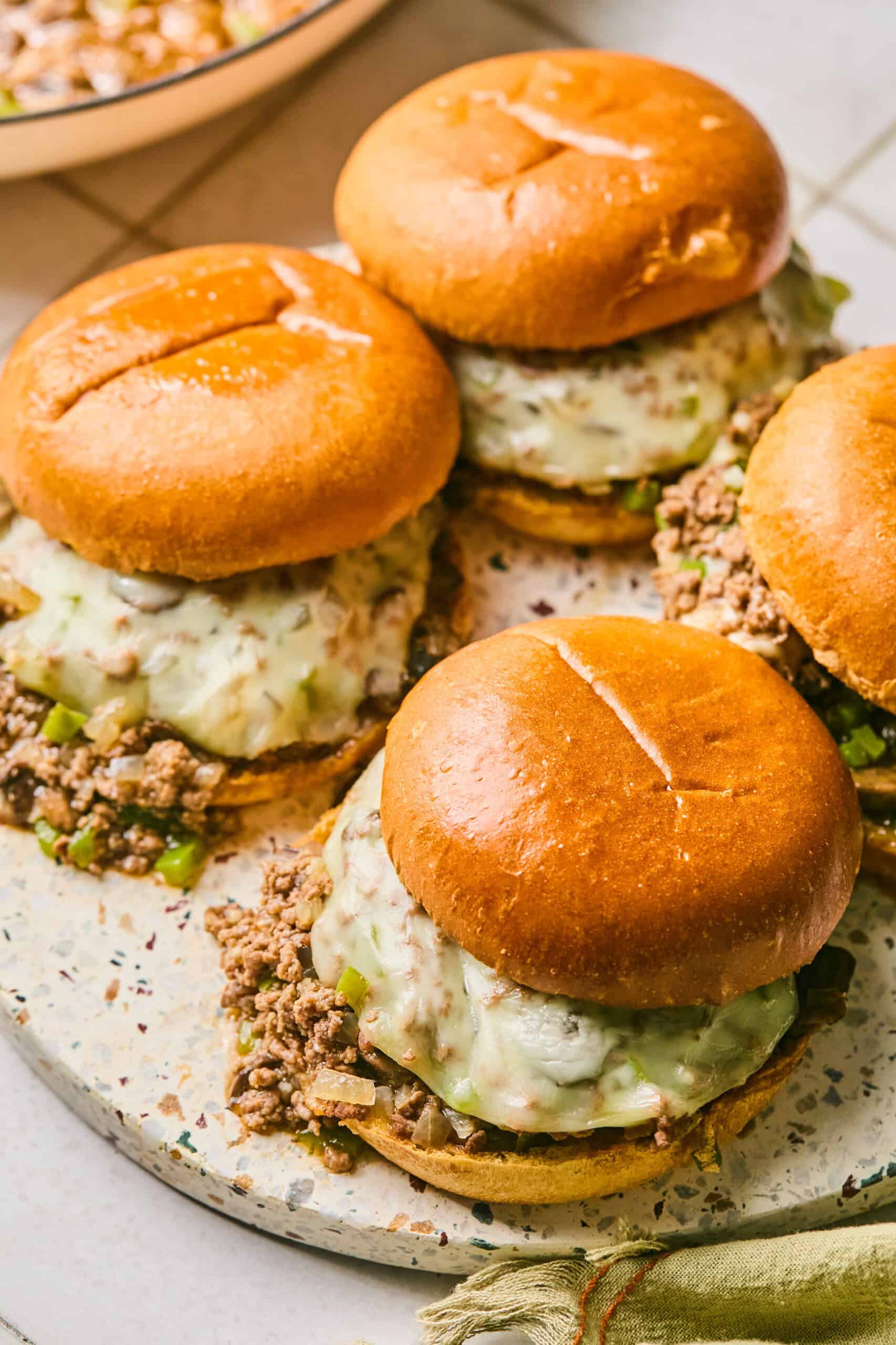 Four burgers with melted cheese and seasoned ground beef on a round platter evoke the essence of Philly Cheesesteak Sloppy Joes. The glossy buns hold a filling with visible pieces of green bell pepper, while a linen napkin peeks out beside the platter.