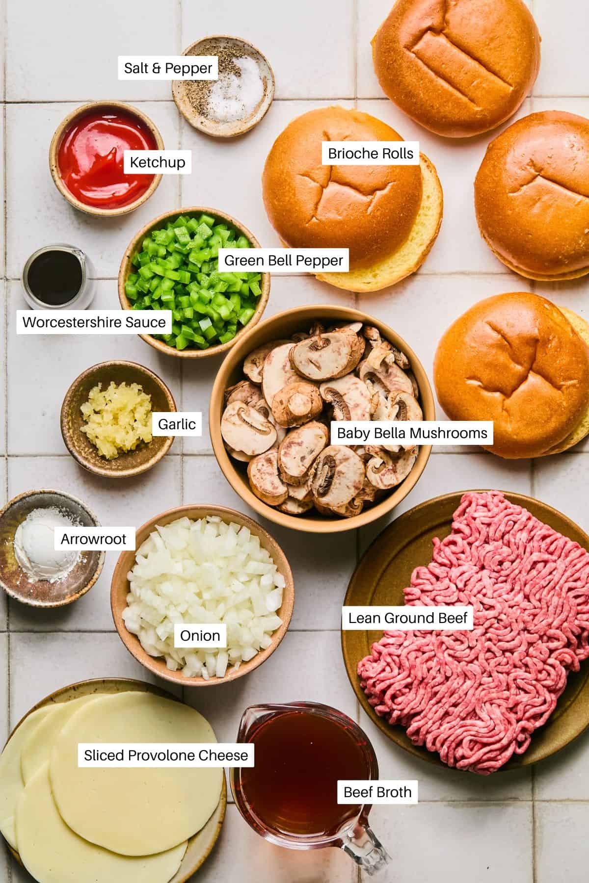 Ingredients for Philly Cheesesteak Sloppy Joes are spread out on a white surface: salt, pepper, ketchup, Worcestershire sauce, arrowroot, diced onion, minced garlic, sliced green bell pepper, baby bella mushrooms, brioche rolls, sliced provolone cheese, ground beef, and beef broth.