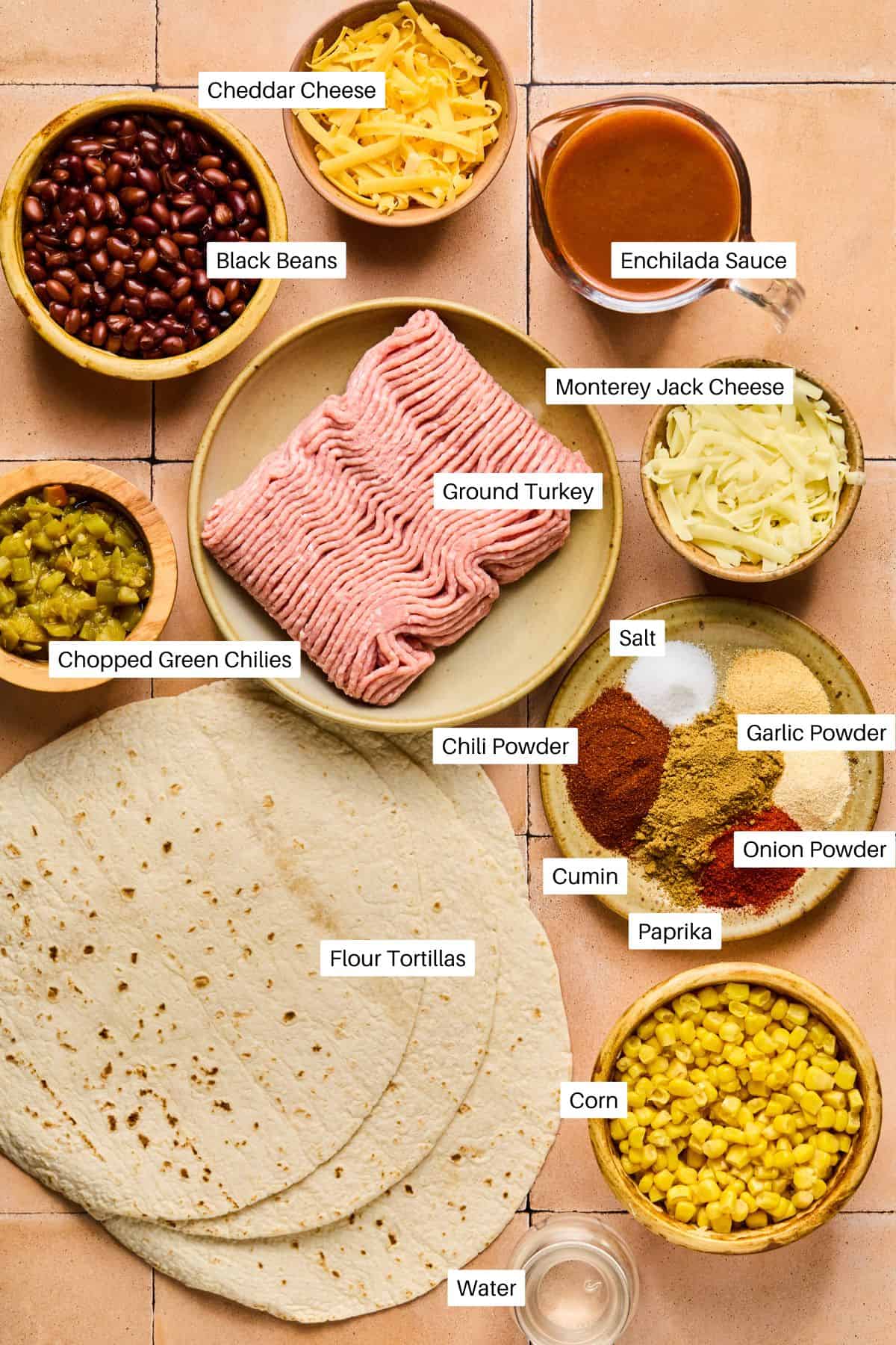 Ingredients for a taco pie-inspired enchilada are spread on a tile surface: flour tortillas, ground turkey, cheddar cheese, Monterey Jack cheese, black beans, enchilada sauce, chopped green chilies, corn, spices, and water.