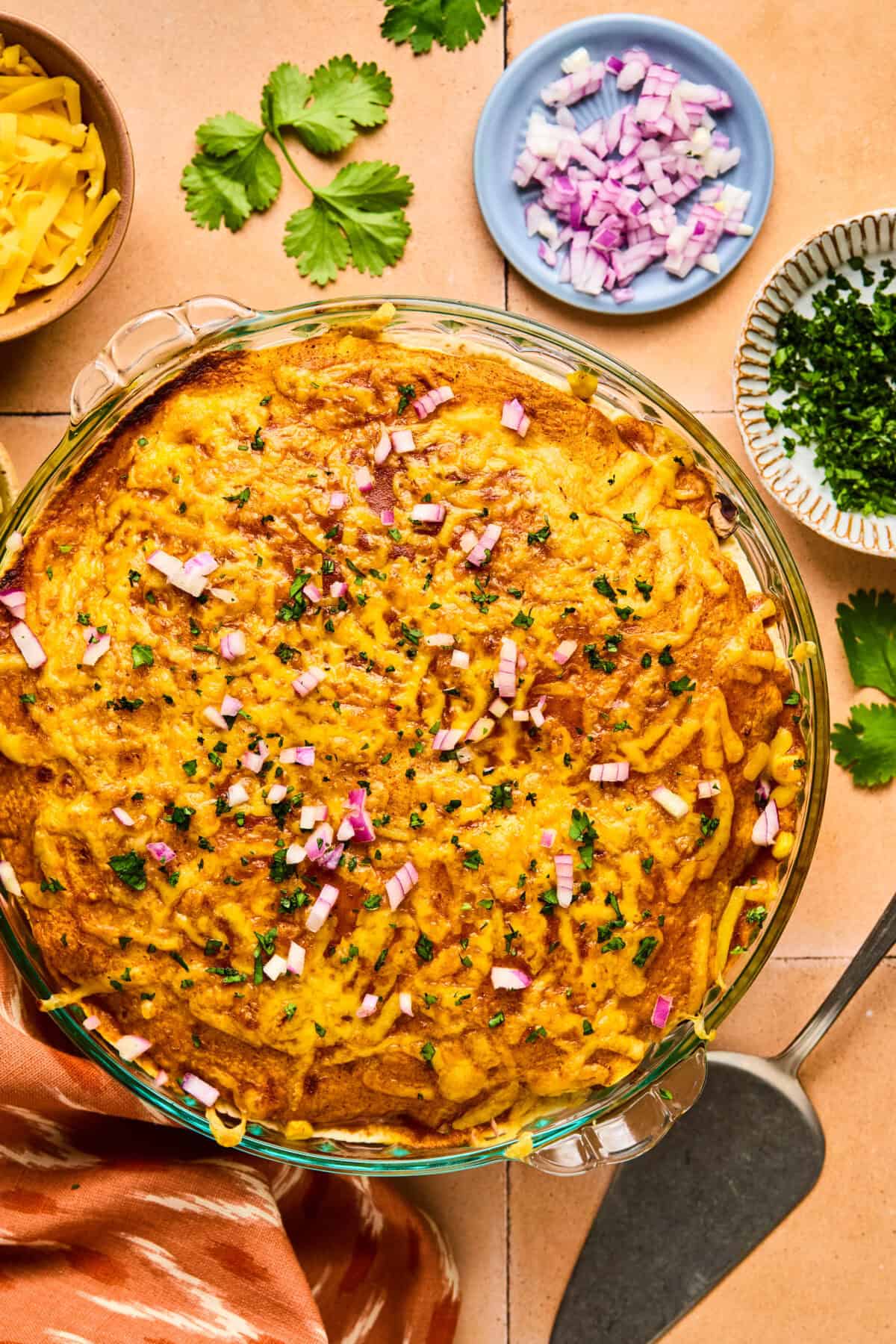 A delicious Taco Pie casserole topped with melted cheese, chopped red onions, and herbs in a glass dish. Surrounding the dish are small bowls of cheese, chopped onions, herbs, and fresh cilantro. A serving spatula rests nearby.