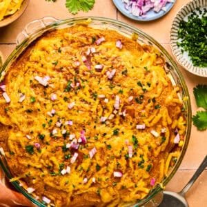 A delicious Taco Pie casserole topped with melted cheese, chopped red onions, and herbs in a glass dish. Surrounding the dish are small bowls of cheese, chopped onions, herbs, and fresh cilantro. A serving spatula rests nearby.