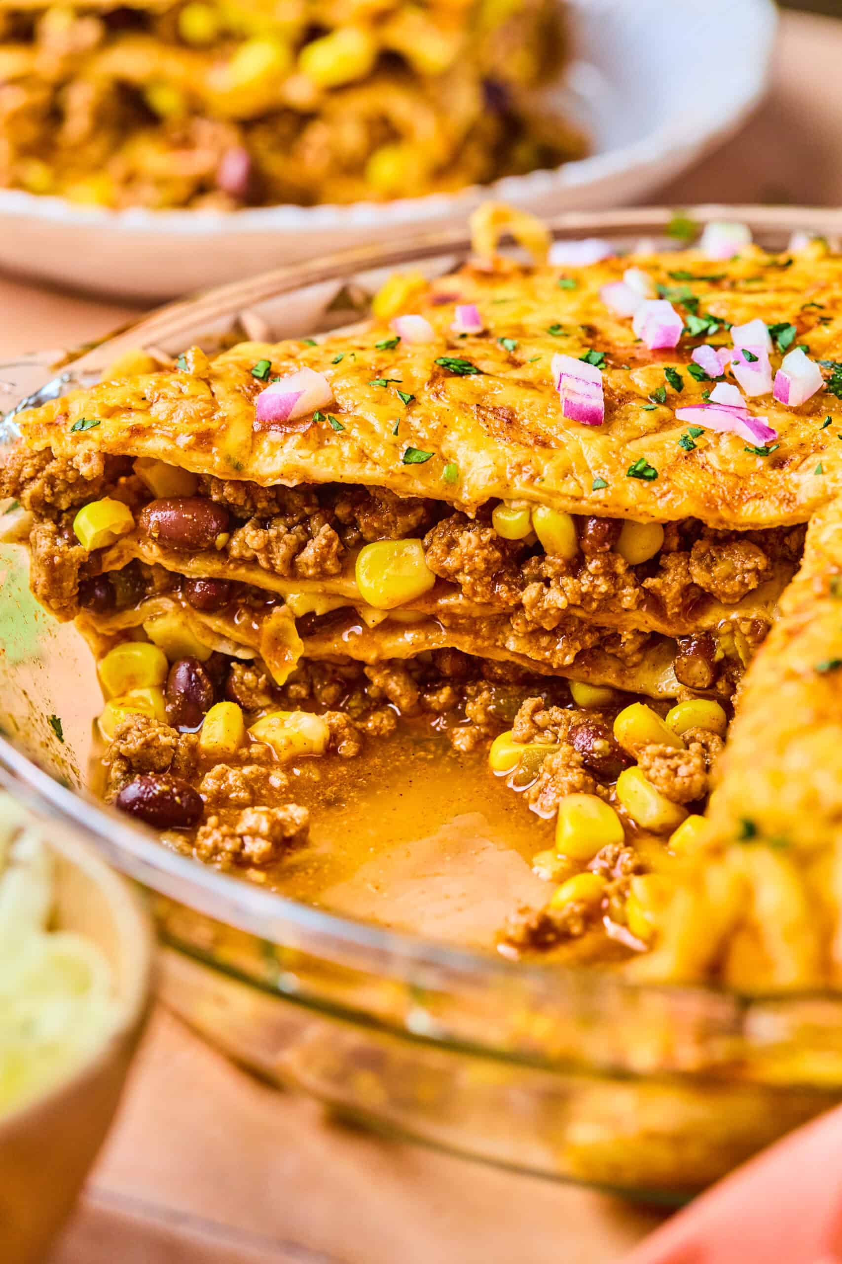 Close-up of a slice of layered taco pie casserole in a glass dish. The dish reveals layers of seasoned ground beef, corn, beans, and cheese, topped with chopped red onions and cilantro. The casserole is set on a wooden surface.