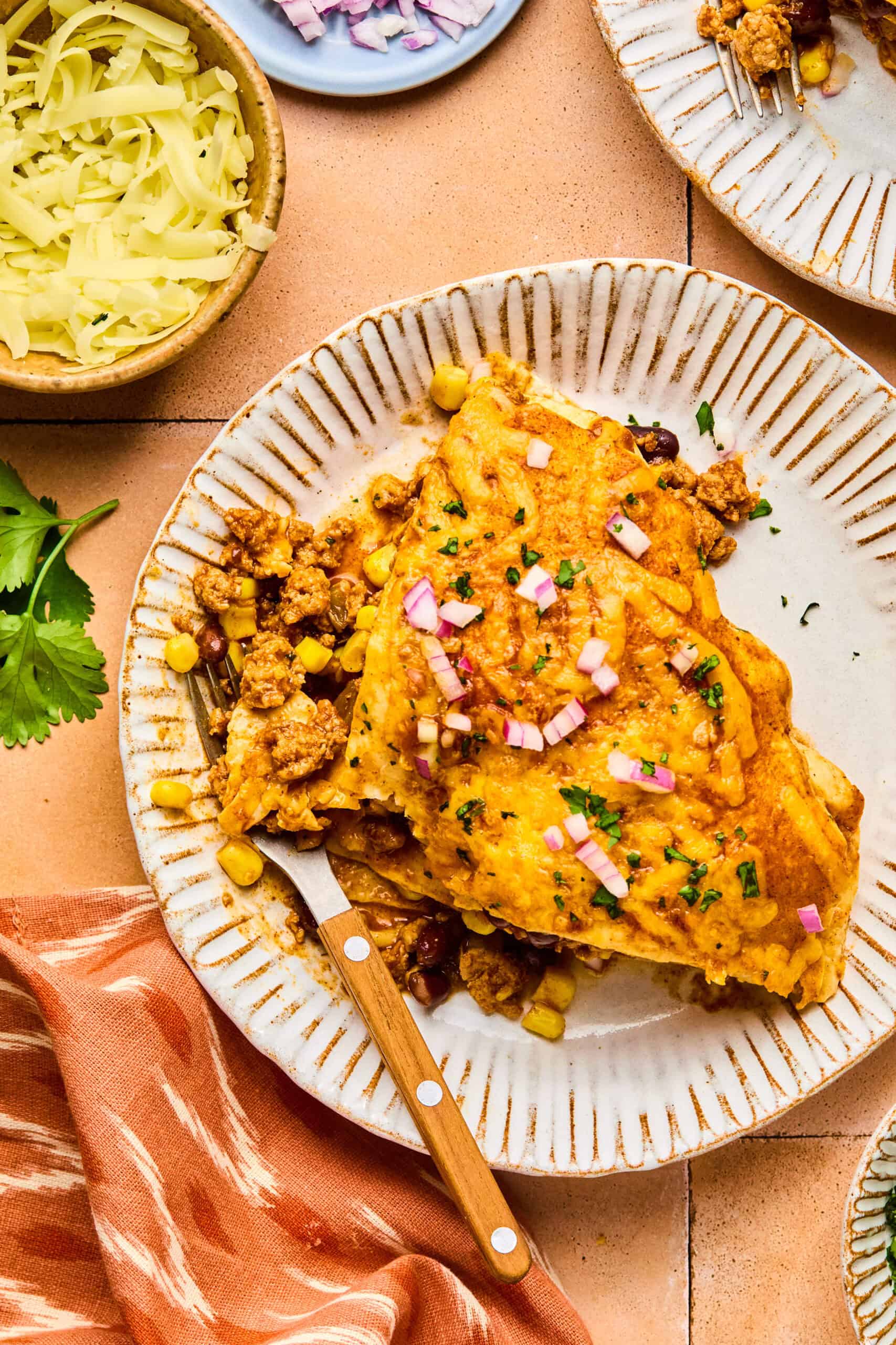 A plate with two cheesy enchiladas topped with chopped onions and cilantro evokes flavors reminiscent of a delightful taco pie. A fork rests on the side, while bowls of shredded cheese, diced red onions, and fresh cilantro surround it. An orange-striped napkin is nearby.