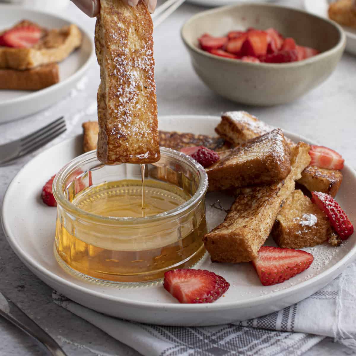 French toast sticks being dipping into maple syrup with strawberries on the side.
