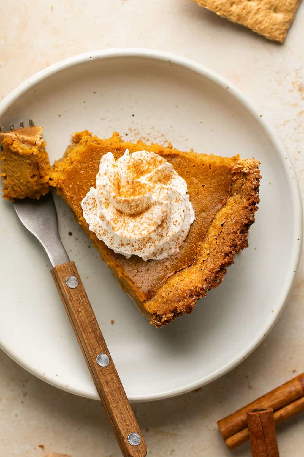 A slice of pumpkin pie with whipped cream and cinnamon on top with a bite taken out of it with a fork.