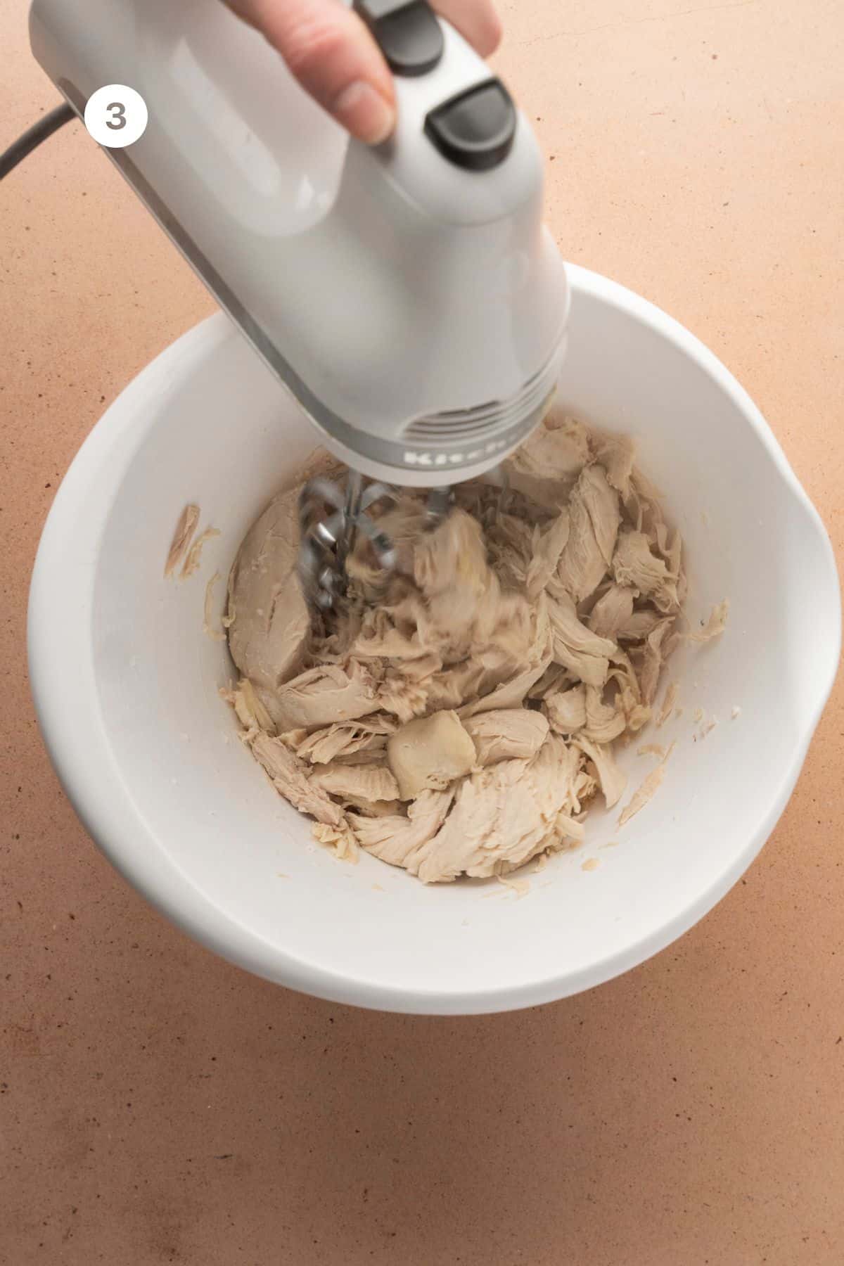 Shredding chicken with a hand mixer in a bowl. 
