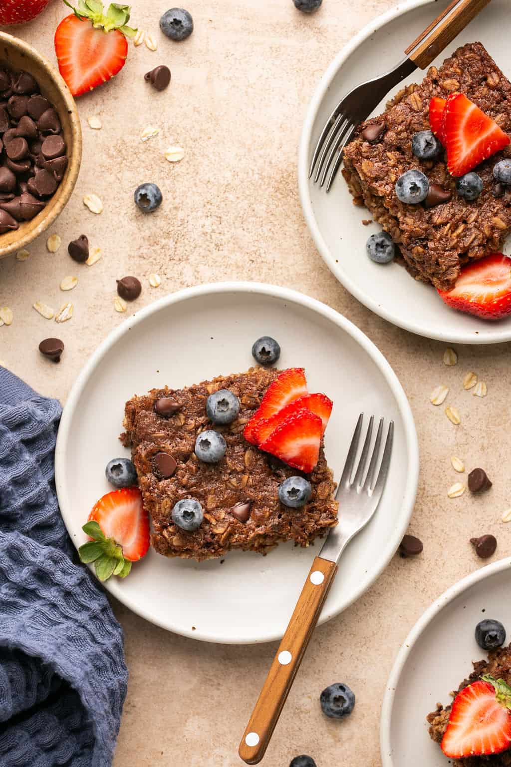 Plates of chocolate baked oats topped with blueberries and sliced strawberries. A wooden-handled fork rests on each plate, and chocolate chips are scattered around. A bowl of chocolate chips and a blue cloth are nearby.