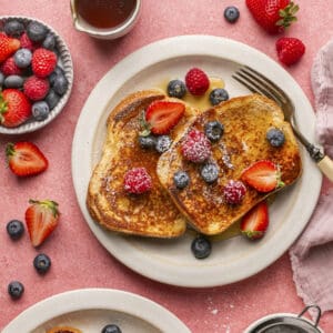 French toast on a plate with berries, powdered sugar and maple syrup on top.