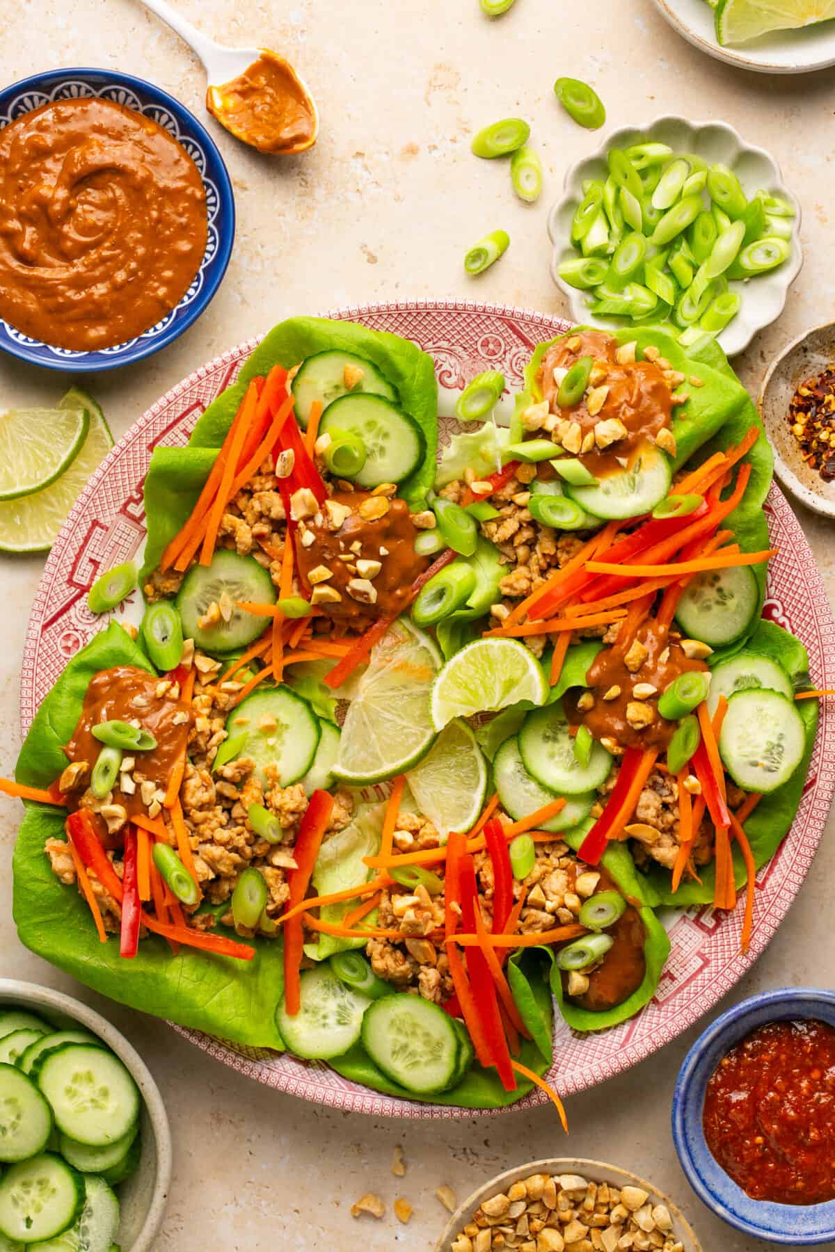 Plate full of assembled lettuce wraps with chicken, cucumbers, bell peppers, carrots, peanuts and extra peanut sauce on top.