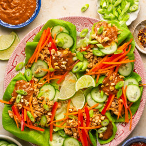 Plate full of assembled lettuce wraps with chicken, cucumbers, bell peppers, carrots, peanuts and extra peanut sauce on top.