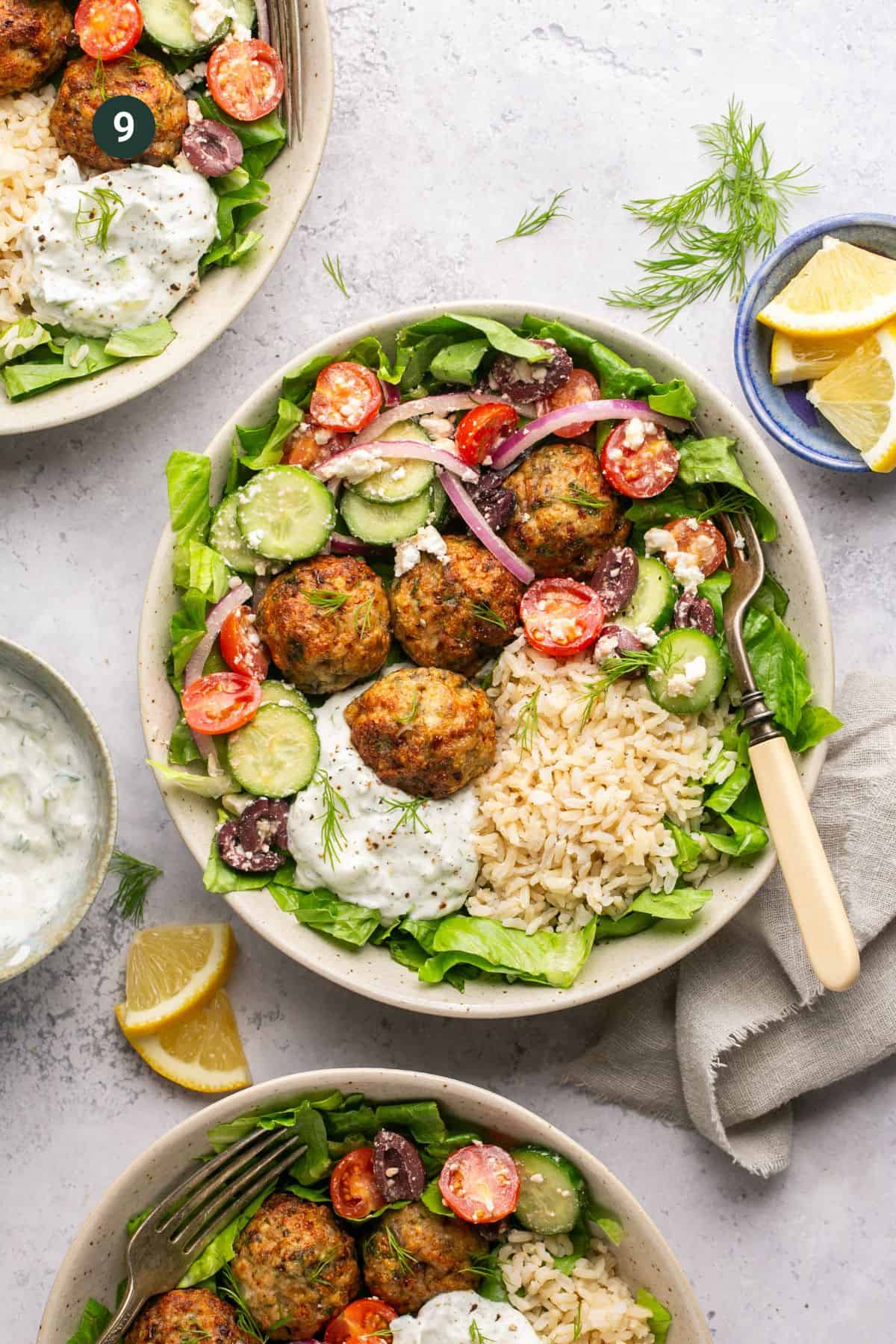 A bowl filled with Mediterranean Chicken Meatballs, falafel, brown rice, sliced cucumbers, cherry tomatoes, red onions, olives, and lettuce is accompanied by a dollop of tzatziki sauce. A small plate of lemon wedges and a cloth napkin with a fork are nearby.