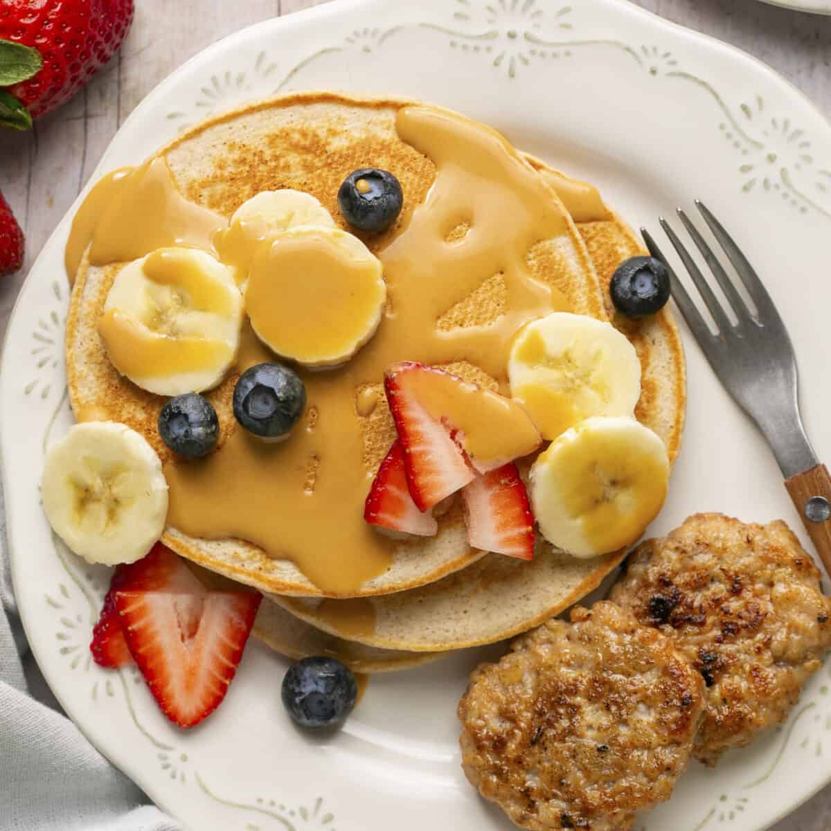 Pancakes on a plate with peanut butter sauce, sliced berries and sliced bananas on top with a side of sausage patties.