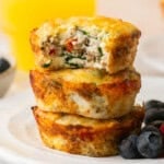 Three stacked egg muffins, akin to egg white bites, rest on a white plate filled with chopped vegetables and sausage. The top muffin has a bite taken out. Nearby, blueberries are scattered around, with a glass of orange juice in the blurred background.