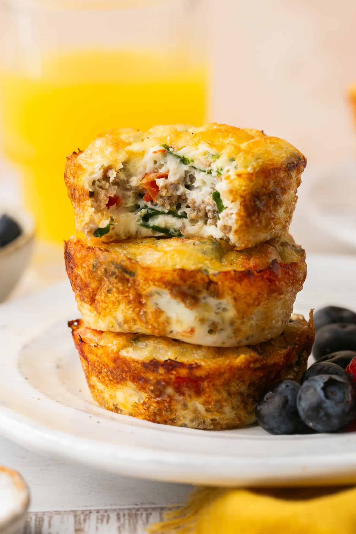 Three stacked egg muffins, akin to egg white bites, rest on a white plate filled with chopped vegetables and sausage. The top muffin has a bite taken out. Nearby, blueberries are scattered around, with a glass of orange juice in the blurred background.