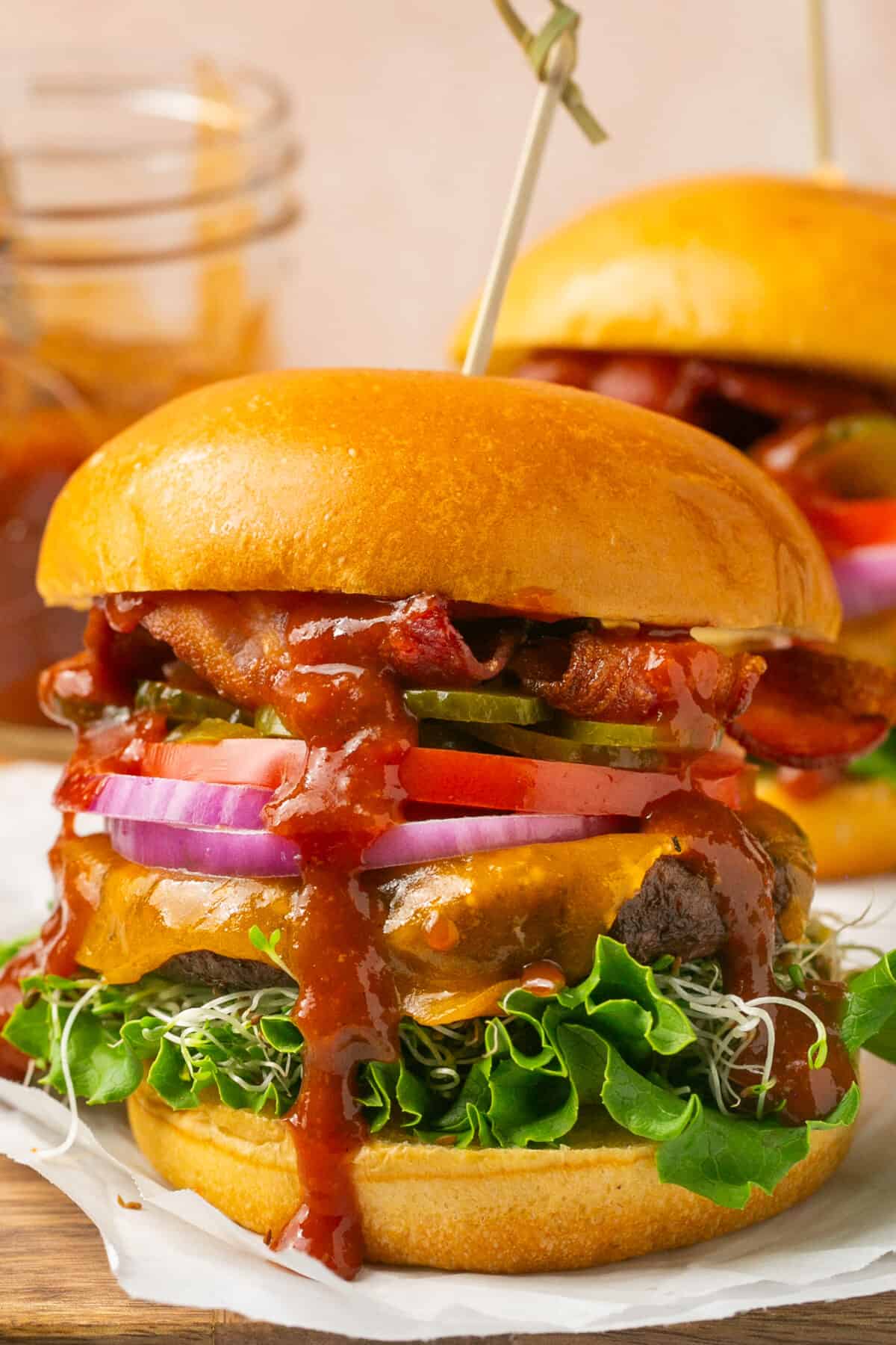 Close up shot of an assembled burgers with lettuce, sprouts, tomato, onion, pickles, cheese burger patties, bacon and homemade bbq sauce on brioche buns.
