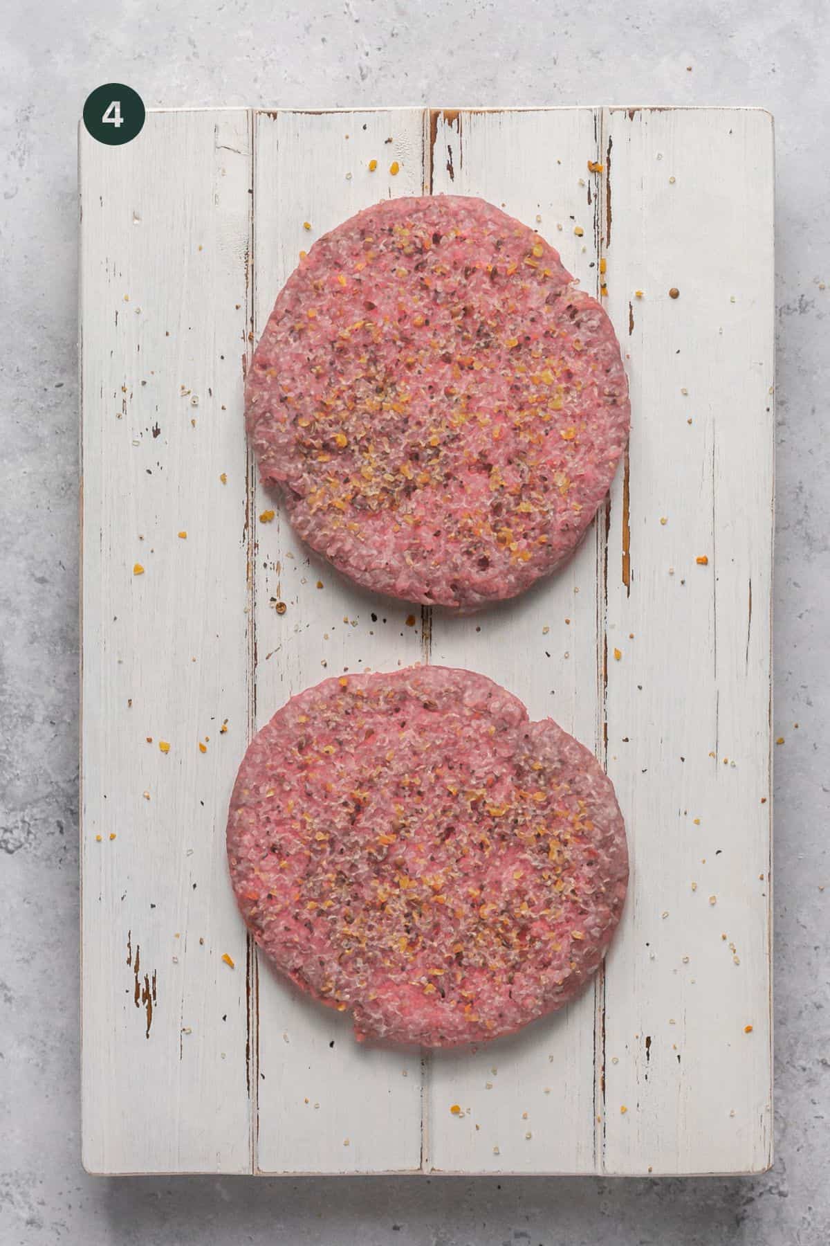 Two frozen burger patties topped with burger seasoning on a board. 