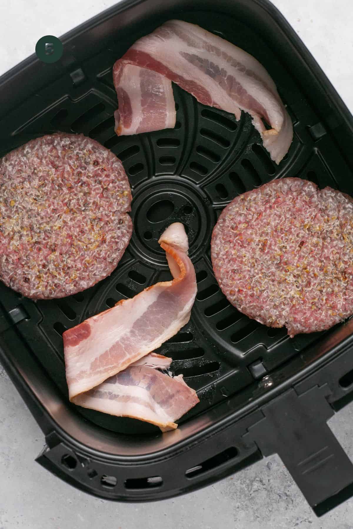 Two frozen burger patties and two bacon strips in the base of the air fryer to air fry.