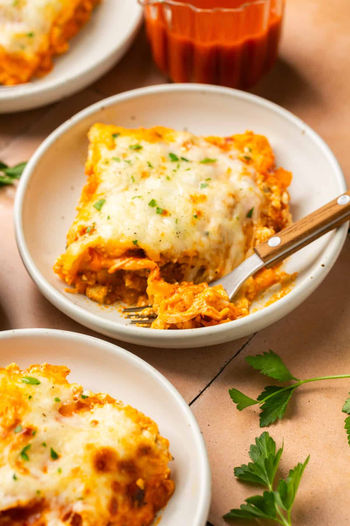 One serving of cheesy lasagna plated with a fork next to another serving and cilantro spread around.