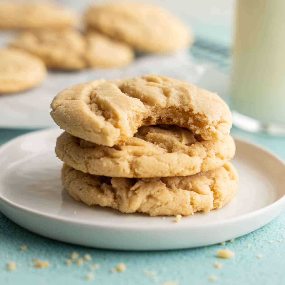 Three cookies stacked on a plate with a bite out of the top one and more cookies behind on a cooling rack.
