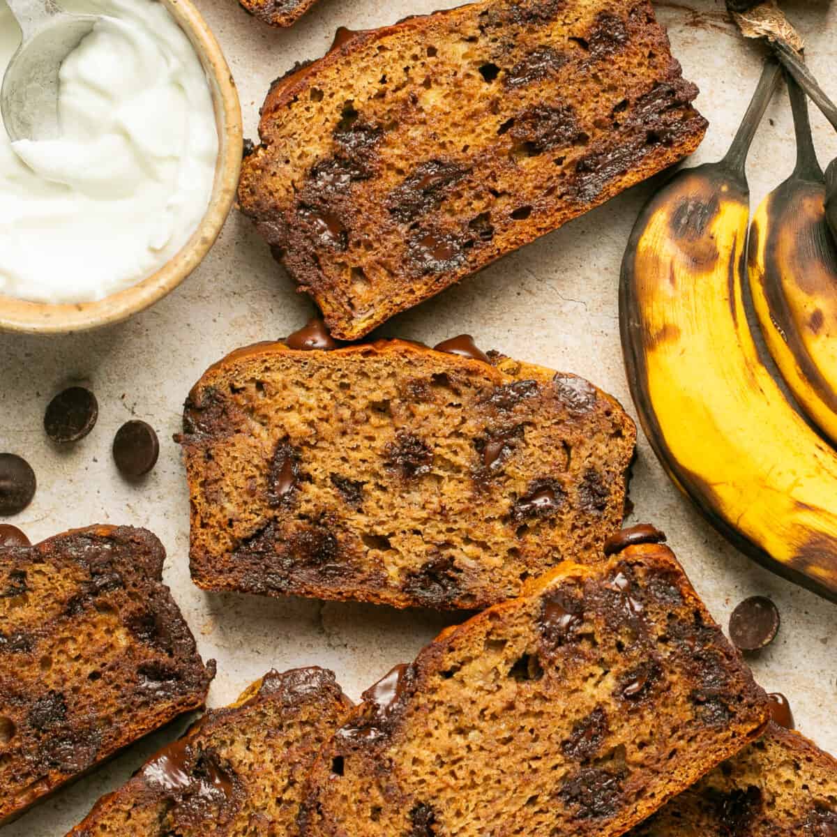 Sliced banana bread with chocolate chips laid out with a small bowl of greek yogurt and a ripe banana.